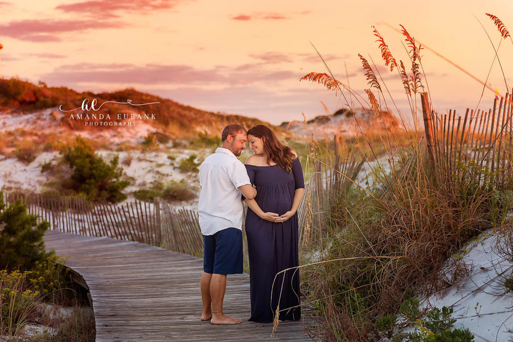 Expecting couple in navy gown and white shirt cradling the baby bump on the dune crossover at sunset in Santa Rosa Beach, Florida.