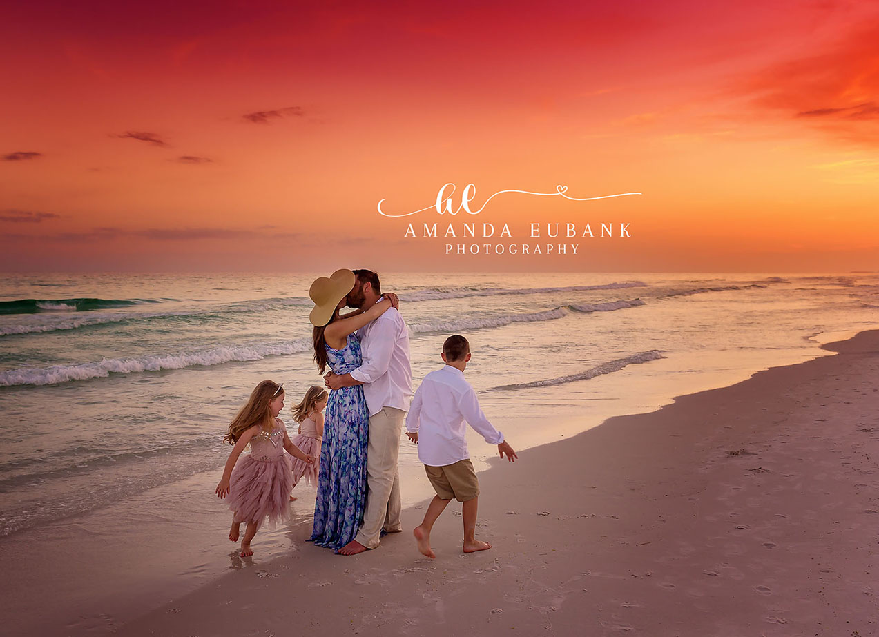 Family of four — mom in a floral dress and a wide sun hat kissing dad in a white linen shirt, with their young daughter in pink and son in white — walking together along the 30A shoreline beneath a vivid coral and orange sunset sky; photographed by Amanda Eubank Photography along Florida’s Emerald Coast.
