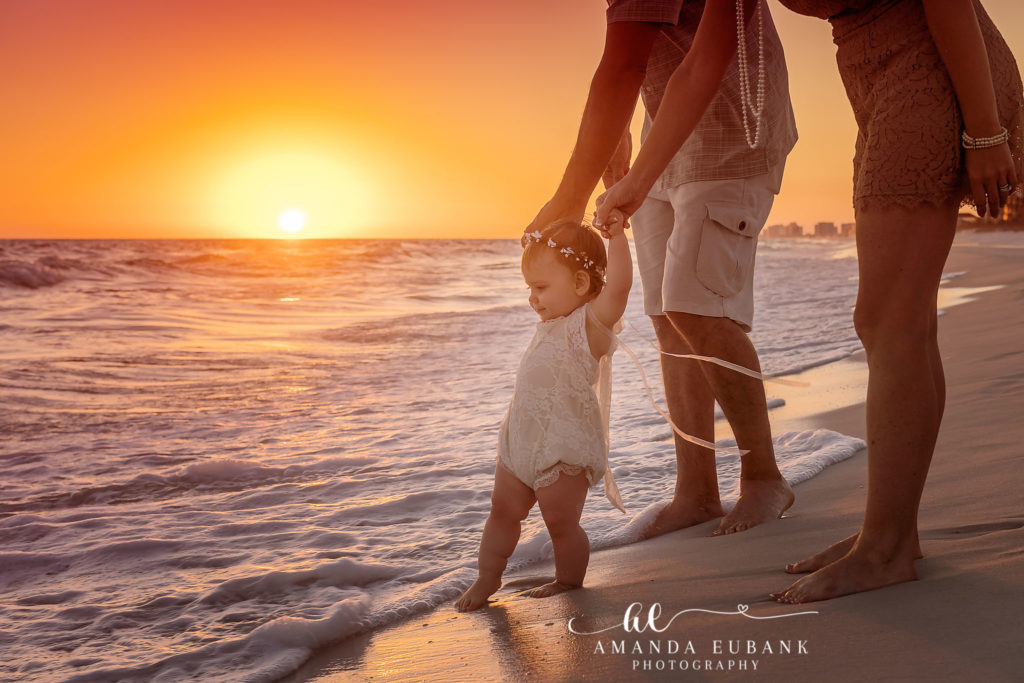 30A first-birthday beach portrait — baby-girl learning to walk between her parents at the shoreline; photographed by Amanda Eubank Photography along Florida’s Emerald Coast.