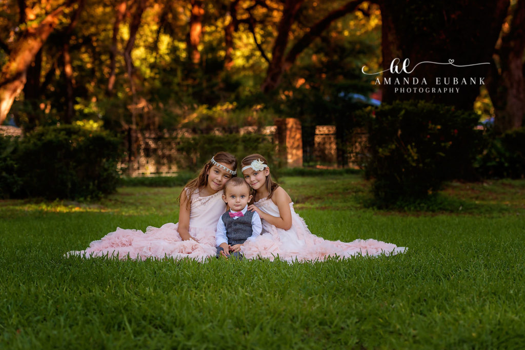 Santa Rosa Beach area portrait of three young siblings — a tender portrait of three young siblings — two older sisters in long blush-pink ruffled tulle gowns flanking a baby boy in a gray vest, white shirt, and pink bow tie — seated on a manicured lawn beneath the golden canopy of live oaks at Eden Gardens State Park; photographed by Amanda Eubank Photography along Florida's Emerald Coast.