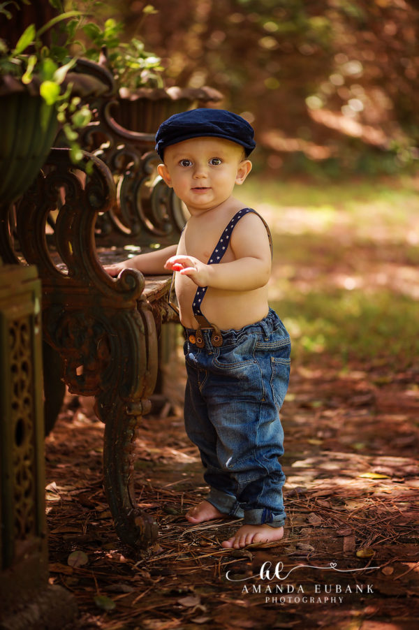 Santa Rosa Beach Area Portrait — standing-by-iron-bench toddler portrait | 30A Family Photographer (Frame 065) Santa Rosa Beach area portrait of toddler boy — the same toddler boy in his navy newsboy cap, navy star-pattern suspenders, and rolled denim jeans standing barefoot on the pine-needle path beside an ornate weathered wrought-iron garden bench in a wooded corner of Eden Gardens; photographed by Amanda Eubank Photography along Florida's Emerald Coast.