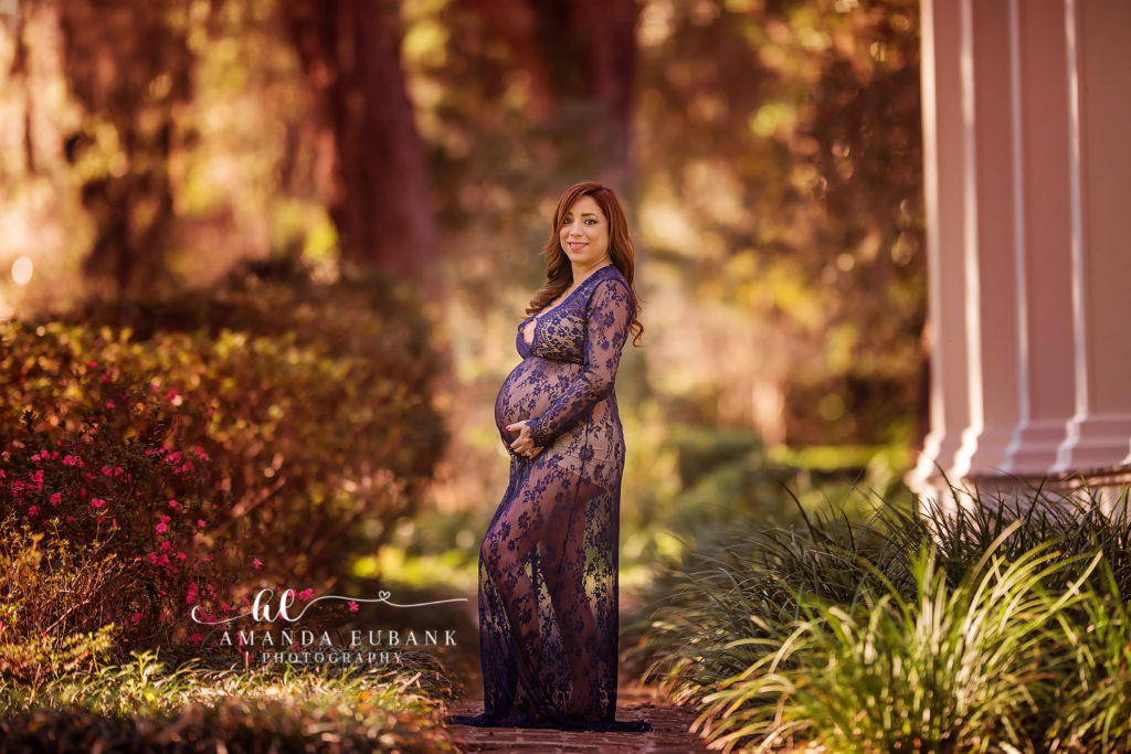 Emerald Coast maternity portrait of expectant mother — standing beside the white classical columns of the Wesley House with magenta azaleas blooming at her feet, hands cradling her bump in her navy floral-lace floor-length maternity gown at Eden Gardens Wesley House column path; a luminous frame photographed by Amanda Eubank Photography along Florida's 30A.