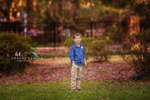 Santa Rosa Beach area family portrait of young son — a young son in a chambray button-down with rolled cuffs and khaki cargo pants standing alone on the autumn-leaf-strewn lawn of Eden Gardens State Park, smiling toward the camera with a wrought-iron fence softly blurred behind him; photographed by Amanda Eubank Photography along Florida's Emerald Coast.
