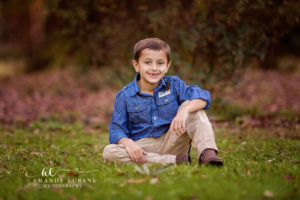 Santa Rosa Beach area family portrait of young son — the same young son in his chambray button-down and khaki cargos seated cross-legged on the grass at Eden Gardens State Park with a soft camellia-pink woodland background blurred behind him; photographed by Amanda Eubank Photography along Florida's Emerald Coast.
