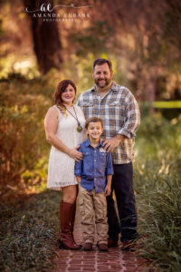 Santa Rosa Beach area family portrait of family of three — a vertical full-length family-of-three portrait standing on the brick path at Eden Gardens State Park — mom in a sleeveless white lace minidress and cognac riding boots, dad in a blue plaid button-down and dark denim, son in a chambray shirt and khakis — with the brick path receding into a soft sun-flared canopy of trees; photographed by Amanda Eubank Photography along Florida's Emerald Coast.