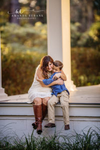 Santa Rosa Beach area family portrait of young son — a portrait of the young son alone seated cross-legged on a sandstone walkway at Eden Gardens with afternoon sun-flare on tropical foliage behind him in his chambray button-down and khakis; photographed by Amanda Eubank Photography along Florida's Emerald Coast.
