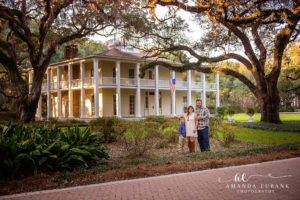 Santa Rosa Beach area family portrait of family of three — a wide architectural family-of-three portrait at the iconic white-columned mansion of Eden Gardens State Park — the historic Wesley House framed by sprawling moss-draped live oaks and an American flag hanging from the second-story balcony — with mom, dad, and son standing together on the brick path in front; photographed by Amanda Eubank Photography along Florida's Emerald Coast.