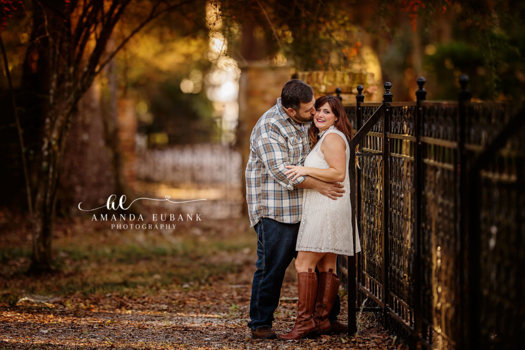 Santa Rosa Beach area family portrait of mom and dad — mom and dad in another moment at the wrought-iron Eden Gardens arbor gate — dad kissing the top of mom's head as she smiles into the camera with her white lace minidress and tall cognac riding boots; photographed by Amanda Eubank Photography along Florida's Emerald Coast.