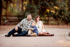 Santa Rosa Beach area family portrait of family of three — a relaxed seated family-of-three portrait on the brick walkway at Eden Gardens with dad in plaid sitting cross-legged on one side, mom in her white lace minidress and cognac boots leaning into him, and the young son in his chambray and khakis tucked between them; photographed by Amanda Eubank Photography along Florida's Emerald Coast.