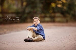 Santa Rosa Beach area family portrait of young son — a smiling close portrait of the young son seated on the brick walkway at Eden Gardens with knees drawn up, in his chambray and khakis, with rich golden bokeh and camellias blurred behind him; photographed by Amanda Eubank Photography along Florida's Emerald Coast.