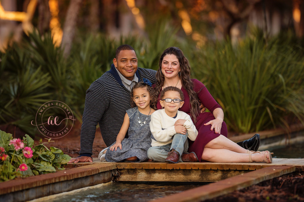 The Rhodes family seated low together on the Watercolor plank bridge, kids snuggled between mom and dad with the water just behind them.