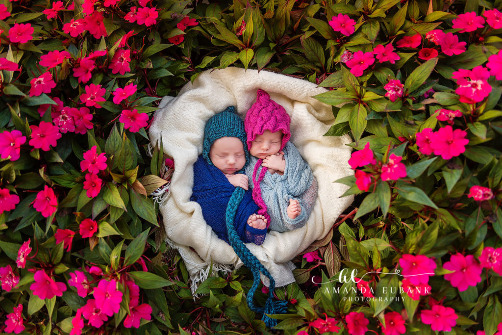 Newborn twins at Watercolor on 30A — the twins sleeping side by side on a cream blanket nestled into a bed of vivid pink impatiens — boy in a teal knit bonnet with a navy wrap, girl in a magenta pixie bonnet with a slate-blue wrap.