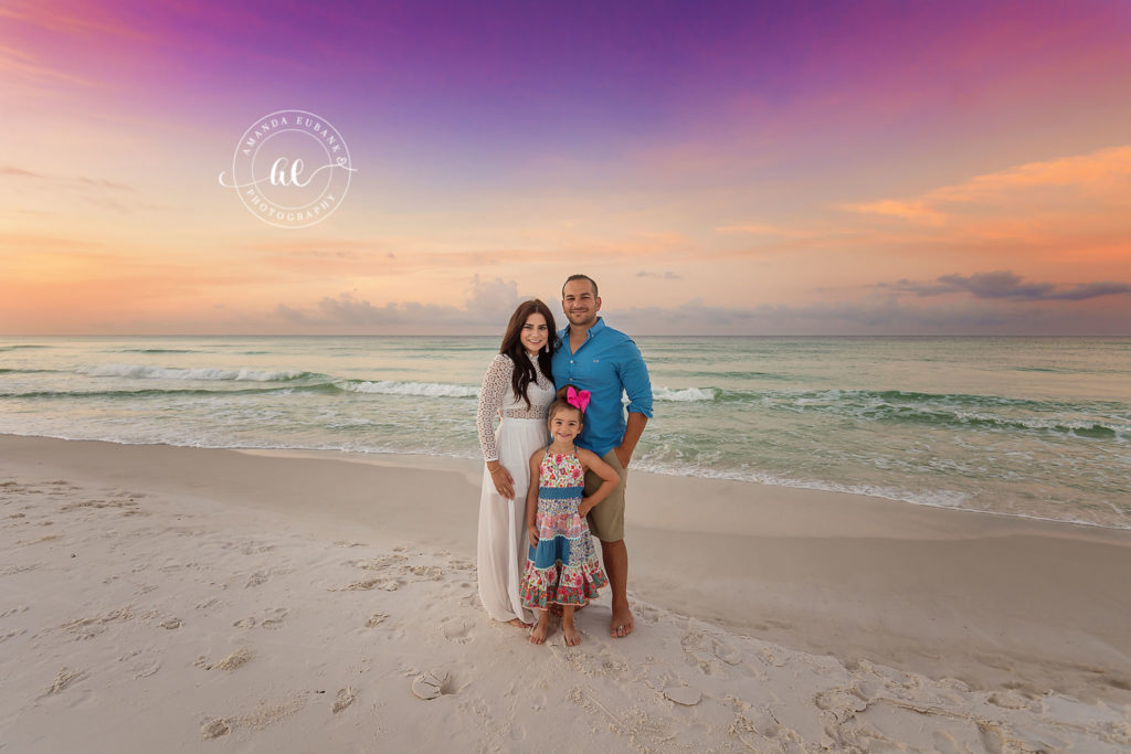 Madi and Casey lifting Aria up high as cotton-candy sunrise color paints the Destin sky.
