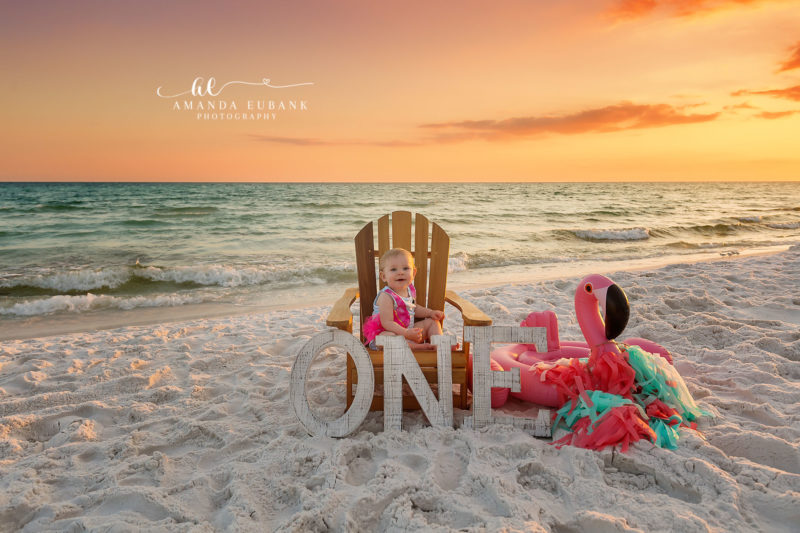 Miramar Beach Florida Baby Photographer — First Birthday Beach Cake Smash Setup Baby Sybi in a wooden Adirondack chair beside her ONE first birthday letters and pink flamingo float at sunset on Miramar Beach.