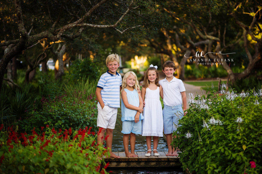 Four cousins (two blondes, two brunettes) standing together in coordinating blues and whites on the wooden plank crossing the Watercolor reflecting fountain on 30A.