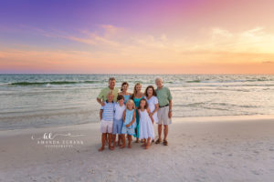 Extended Steinbauer family of ten gathered close on the white sand beach at Watercolor on 30A in a cluster pose under a vivid pink-purple sunset.