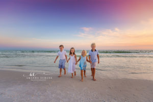 All four Steinbauer cousins walking toward camera hand-in-hand at the surf line at Watercolor on 30A under a soft lavender-and-pink twilight sky.