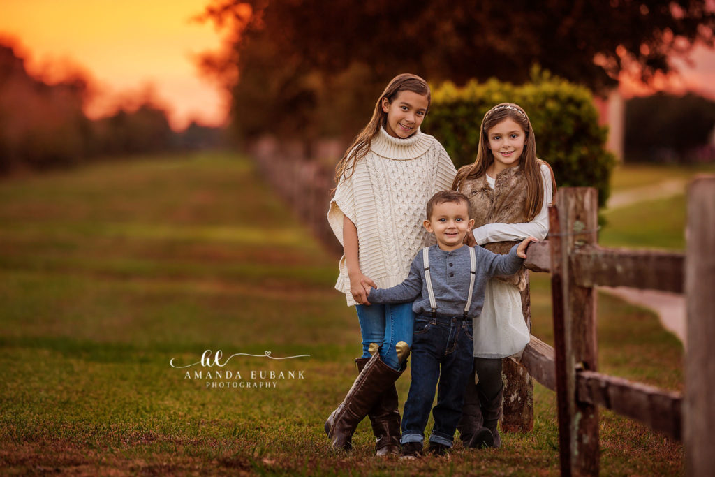 the older sister in a cream cable poncho posed by the field during a Friendswood family autumn session, photographed by Amanda Eubank Photography in soft autumn-park afterglow with wide environmental composition.