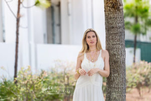 Luminous editorial brand portrait of a model in a cream slip dress with layered gold necklaces leaning on a palm tree captured under sun-bleached open shade
