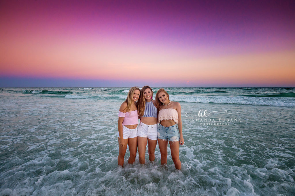the trio sitting together on the white sand at vivid Destin sunset during a sunset Destin sibling trio session on Destin Beach under a multi-toned rainbow sunset, photographed by Amanda Eubank Photography in blue-hour twilight with backlit silhouette.