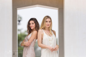 the duo posing with hands behind their heads under the arch during a Texas senior pair-of-friends portrait on the Alys Beach archway, photographed by Amanda Eubank Photography in blue-hour twilight with backlit silhouette.