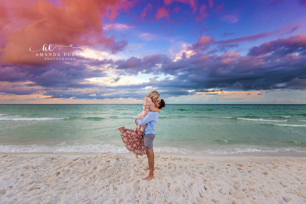 Featured engagement portrait of a fiancé lifting his bride-to-be in a flowing pink floral dress for a kiss above the Destin shoreline