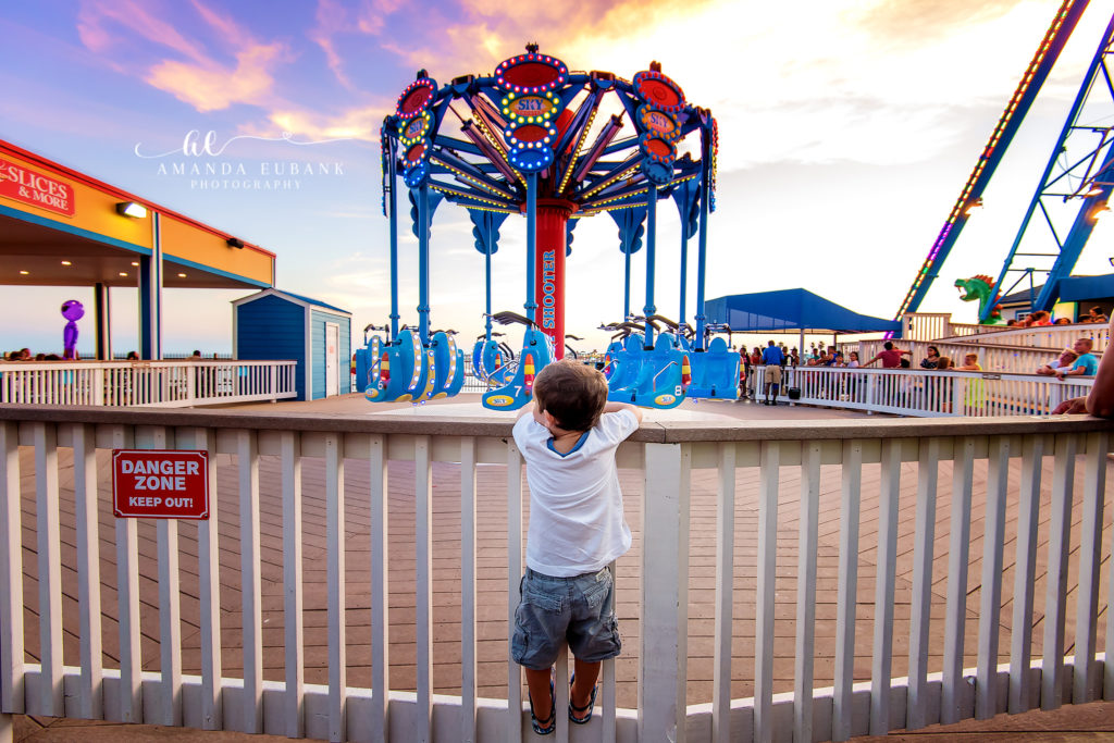 Documentary family-day photograph showing a small spectator leaning on the railing as the Sky Shooter twinkles at sunset during a Galveston Island Pleasure Pier visit