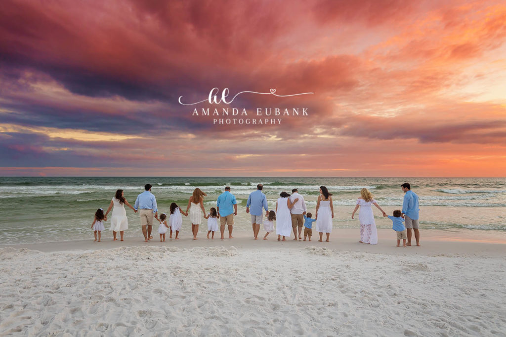 Large extended family in white and blue walking hand-in-hand toward the surf at a fiery pink sunset in Santa Rosa Beach, Florida.