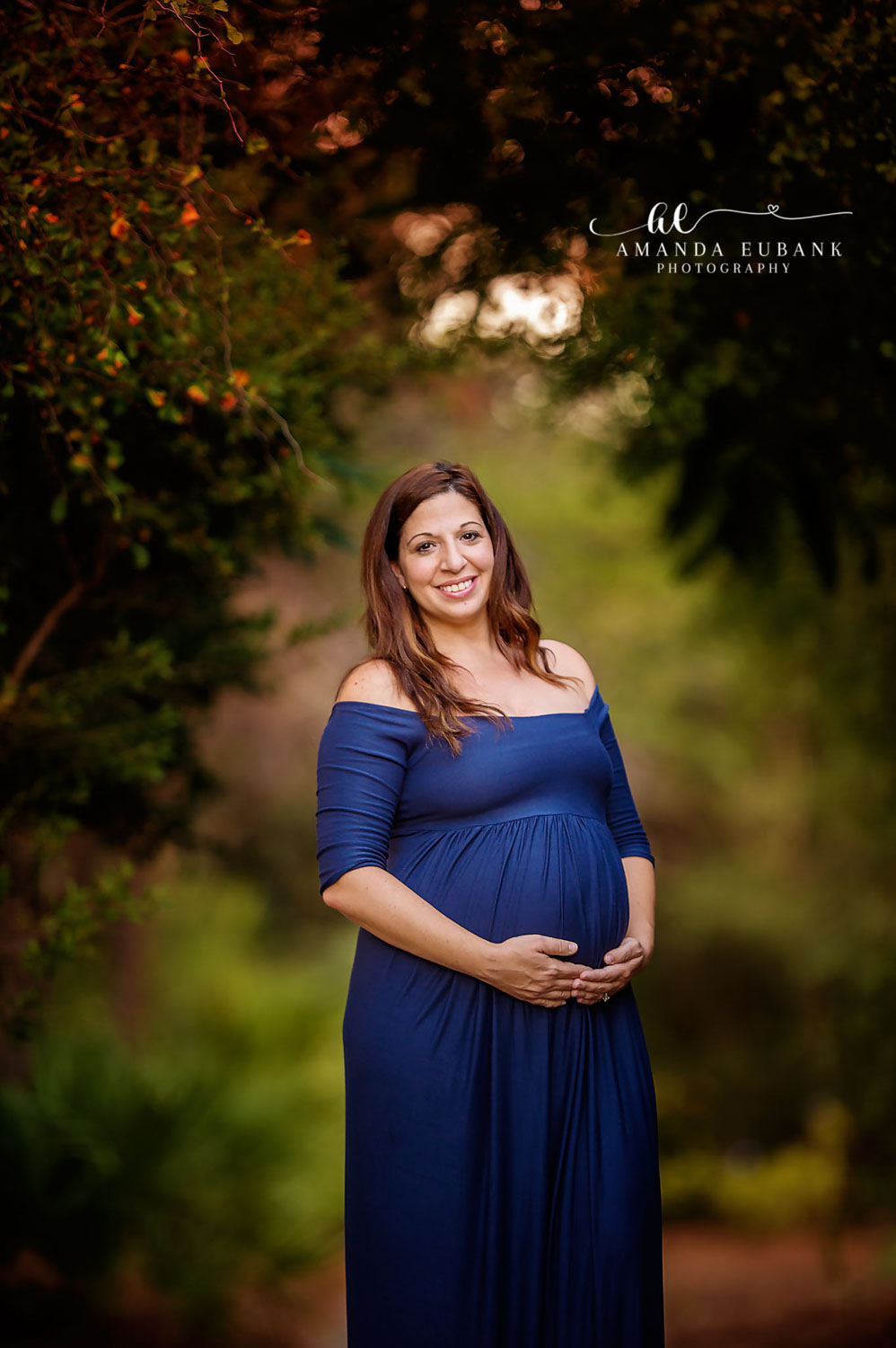 CARILLON BEACH FLORIDA MATERNITY PHOTOGRAPHER Brunette mom-to-be in a royal blue off-shoulder gown cradling her belly in a lush green garden in Carillon Beach, Florida.