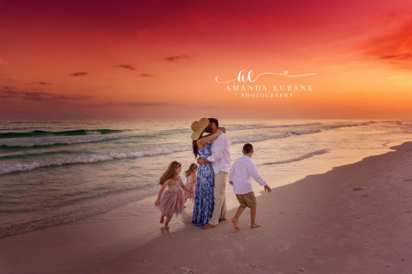 FAMILY PHOTOGRAPHERS IN SANDESTIN FLORIDA Family of five with parents kissing under a sun hat as kids walk beside them on the shoreline at a fiery red sunset on Sandestin Beach, Florida.