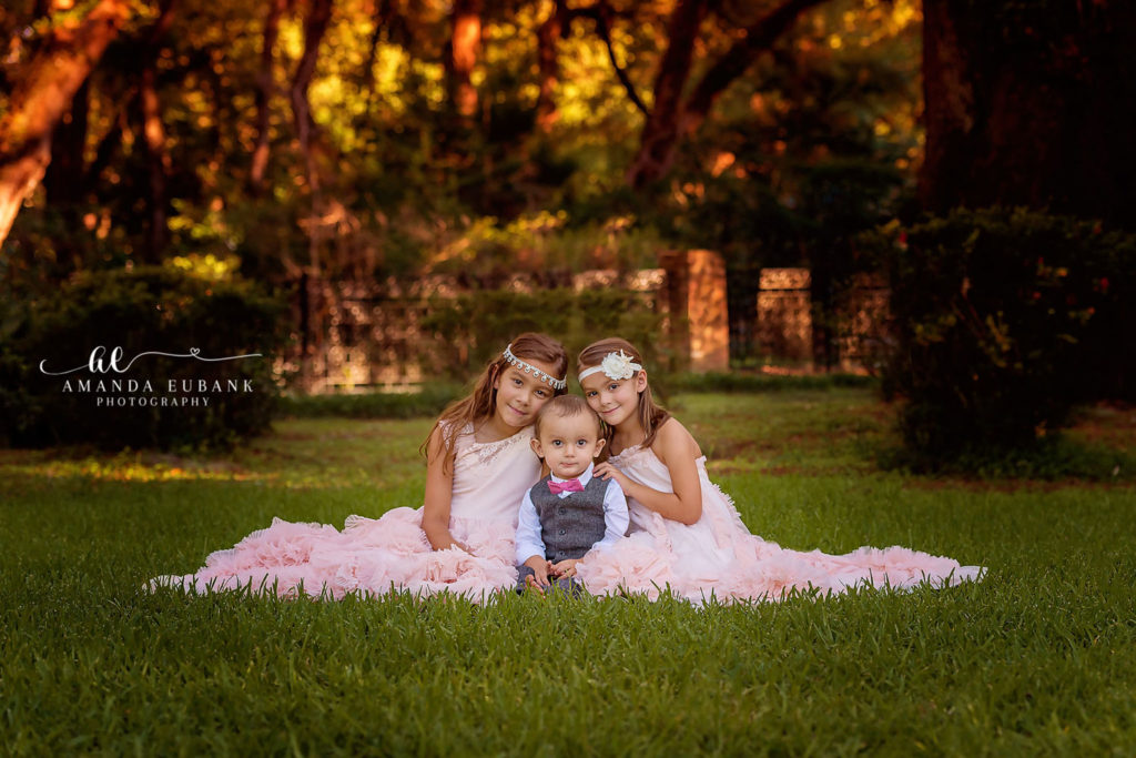 Three siblings — two sisters in blush pink tulle dresses and their little brother in a gray vest and pink bow tie — sitting in the grass at golden hour in Inlet Beach, Florida.