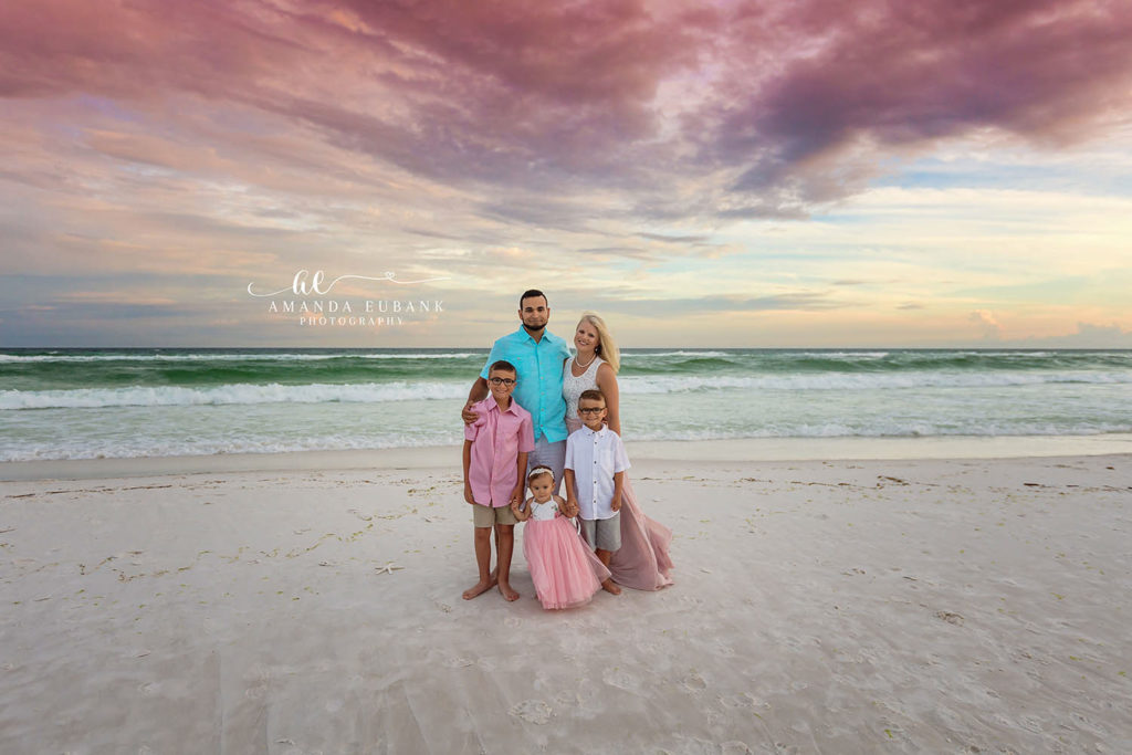 Family of five in coordinated pink, blue, and turquoise posing on the white sand beneath a purple sunset sky on Miramar Beach, Florida.