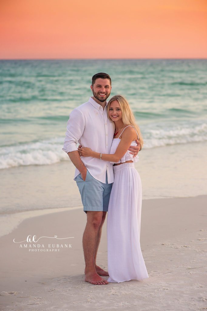 A romantic couple standing on the shoreline of a Destin, Florida beach at sunset, with a vibrant orange and yellow sky reflecting on the Gulf water.