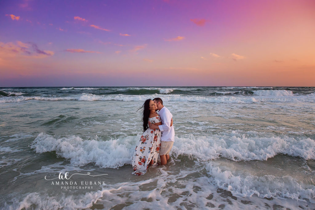 Shannon and Bryan kissing in the ocean surf at sunset with a dramatic purple and orange sky