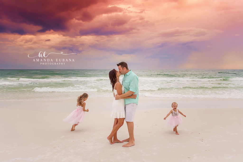 Peccon parents kissing on Santa Rosa Beach with their two daughters in pink tutus at sunset