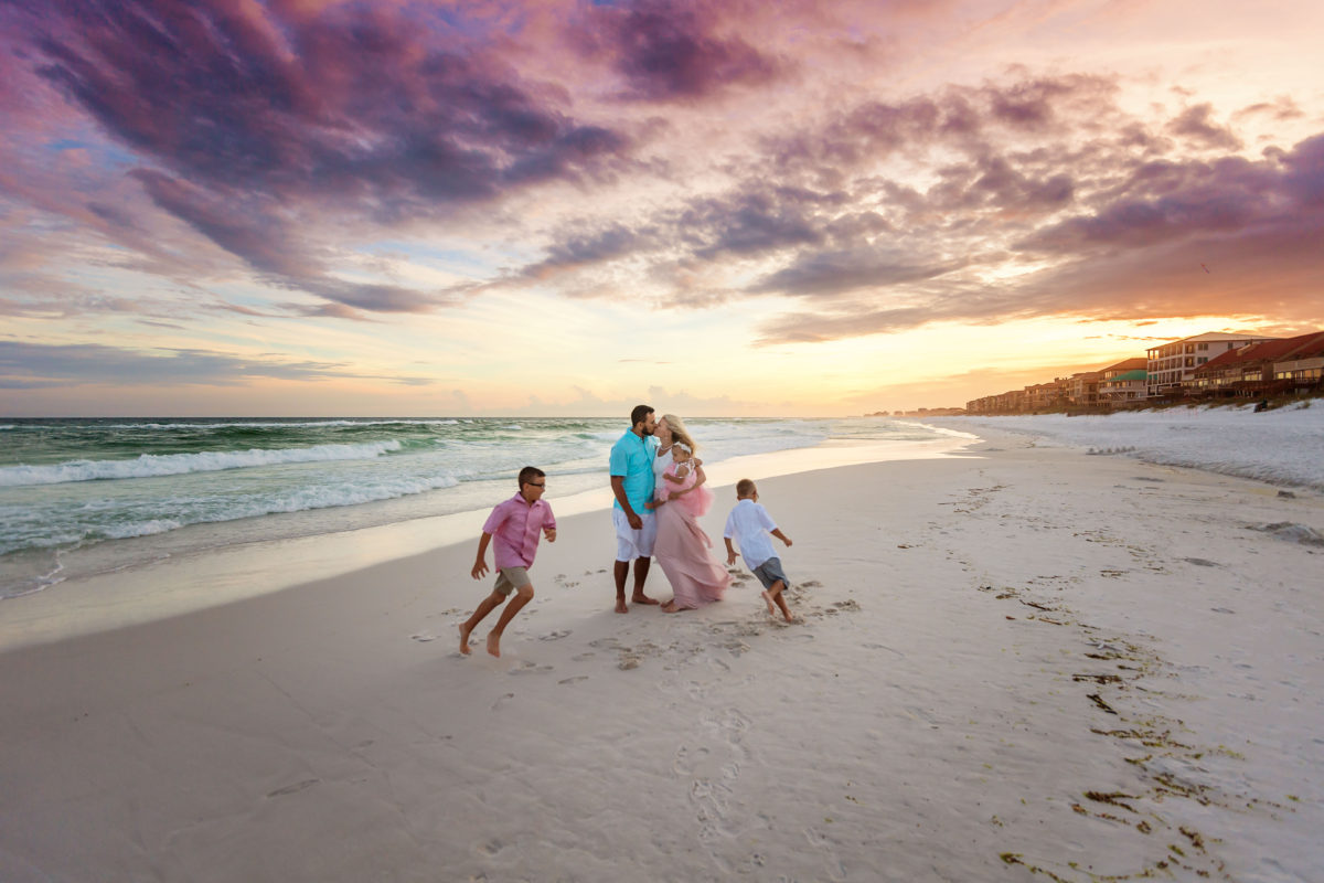 Sandestin Florida Family Photographer — Parents Kiss as Boys Play at Sunset Parents kissing as their two young sons run around them on the shoreline at a purple and orange sunset on Sandestin Beach, Florida.
