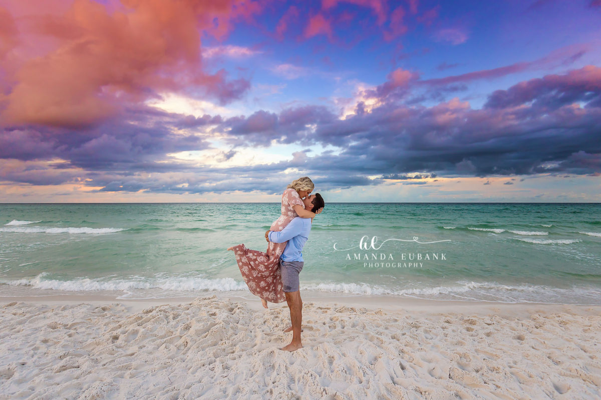 Santa Rosa Beach Sunset Portrait Session A young woman in a flowing dress walking along the shoreline of Santa Rosa Beach under a dramatic sunset with purple and orange clouds.