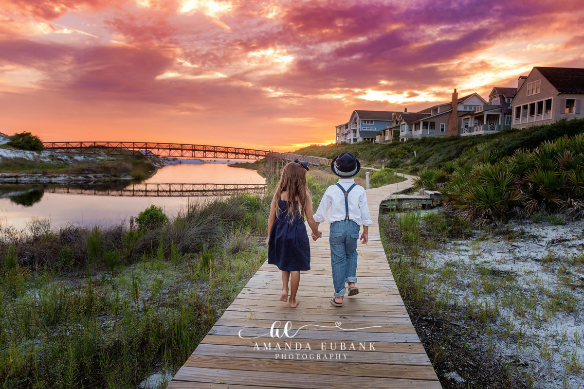 Watersound Florida Family Photographer — Brother and Sister on the Dune Lake Boardwalk at Sunset Young girl in a navy dress and boy in a hat and suspenders walking hand-in-hand along the wooden boardwalk over the dune lake at sunset in Watersound, Florida.