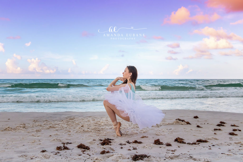 Ballerina in deep grand plié with hand to chin and tutu cascading along the beach at sunrise