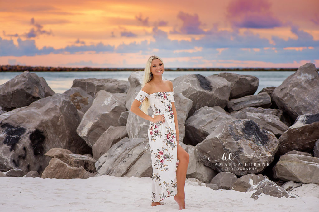 Senior girl in a white off-shoulder floral maxi dress with a high slit standing on white sand beside the rock jetty under a vivid orange and pink sunset