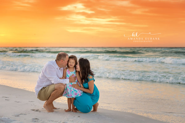 Miramar Beach Sunrise Family Cheek Kisses for Toddler Daughter | Destin Family Photographer Mom and dad kissing their daughter's cheeks while crouched in the sand at fiery sunrise