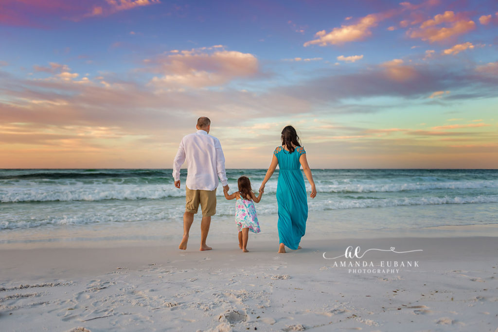 The Elliott family walking hand-in-hand toward the emerald Gulf beneath a soft mauve dawn sky