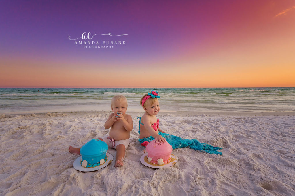 Both twins side by side mid-cake-smash beneath a dramatic sunset sky