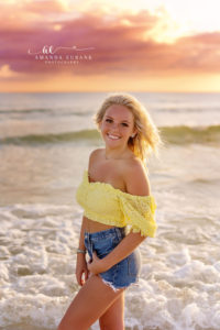 Ali in a yellow off-shoulder eyelet top and denim shorts seated on the 30A sand at sunset.