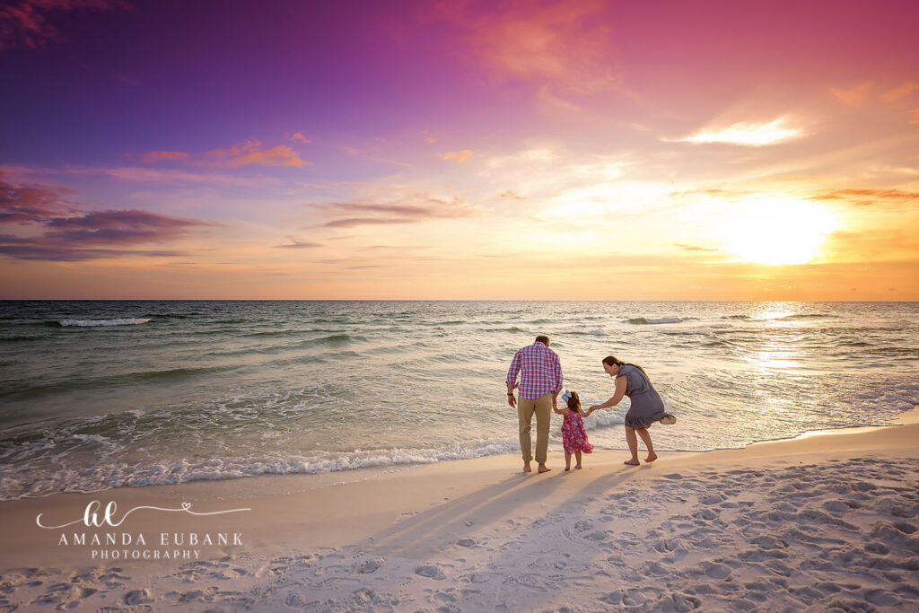 Z parents holding their daughter at the Gulf shoreline beneath a magenta sky