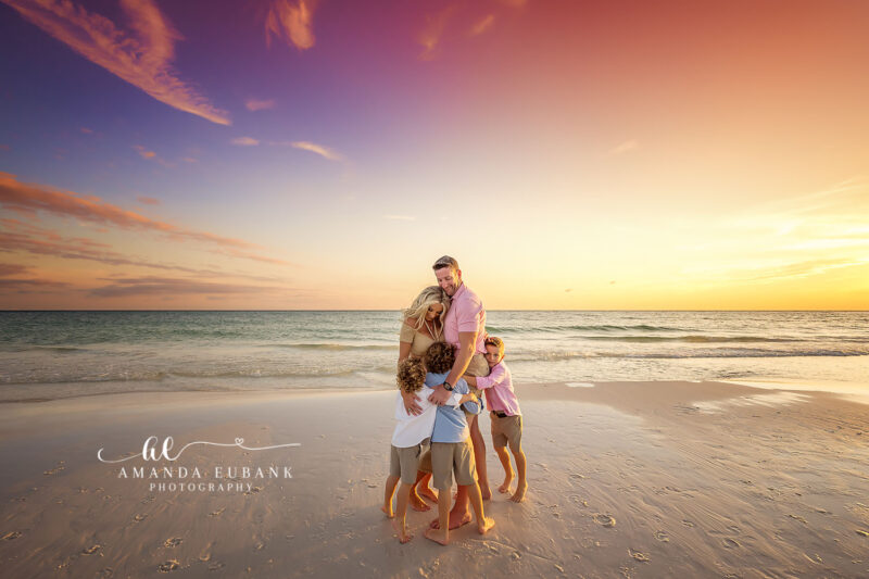 Parents and three young sons share a tight beachside huddle as a violet and peach sunset glows over the Gulf in Miramar Beach, Florida.