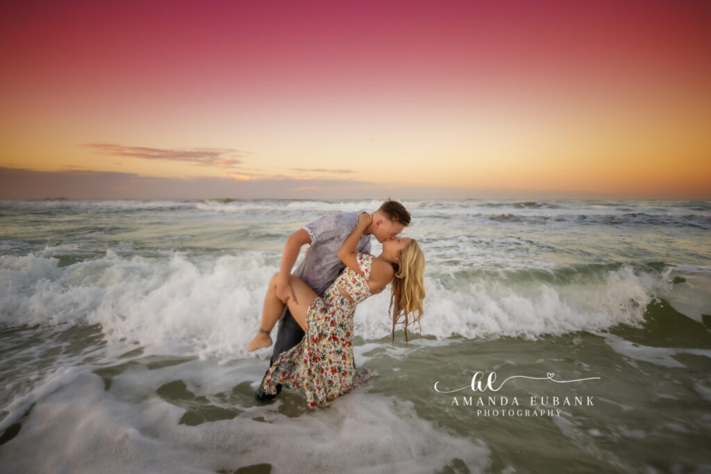 Young couple — guy in a soft grey button-down with denim, blonde girl in an off-shoulder white floral maxi dress — hand in hand by the surf along 30A's Gulf coast at gentle pastel afterglow.
