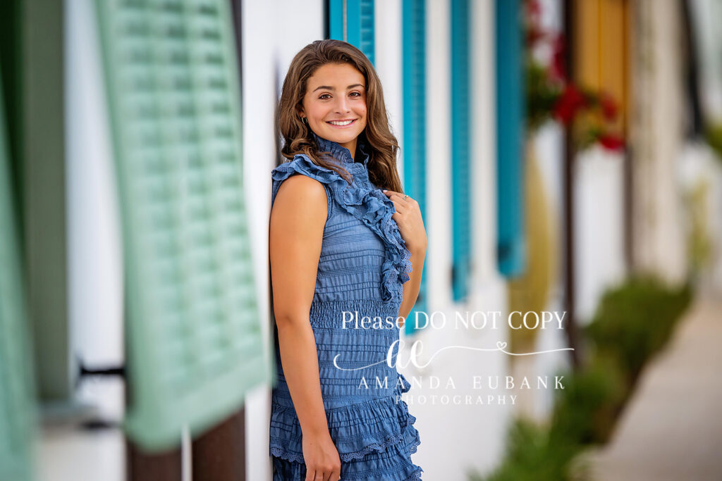 Brunette senior in a cornflower-blue ruffled smocked mini-dress with neutral block-heel sandals, leaning against a sleek white architectural balcony along Alys Beach for an Alys Beach senior portrait session.