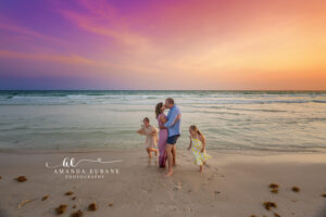 Two young sisters — older girl in a soft beige eyelet tassel dress, younger sister in a white lemon-print sundress — crouching to inspect tiny shells during a Rosemary Beach sunset family session.