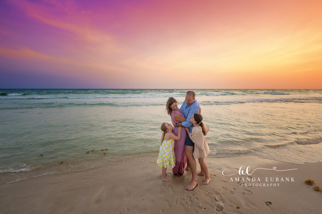 Two young sisters — older girl in a soft beige eyelet tassel dress, younger sister in a white lemon-print sundress — silhouetted against the horizon during a Rosemary Beach sunset family session.